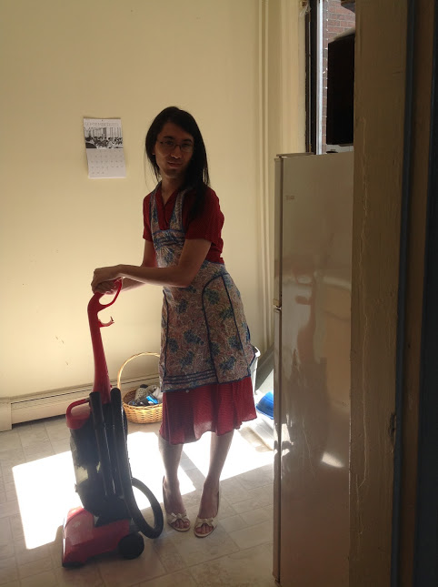 Marina wears an ironically 1950s housewife-esque outfit while pretending to vacuum.